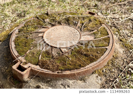 A rusty iron hatch with a pattern covered moss above an observation well is located next to a dry grass in the daytime. Close-up 113499263