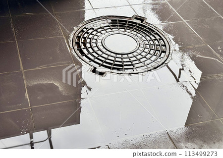A iron hatch with a pattern and covered water above an observation well is located on pedestrian road in the daytime. Close-up 113499273