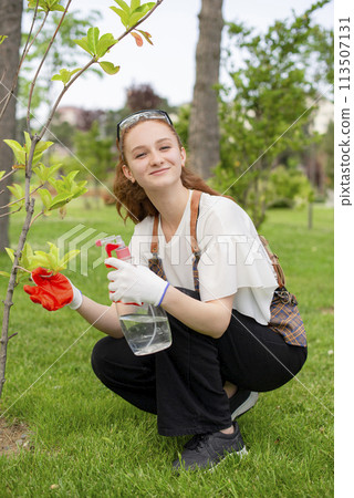 Front view of girl spraying trees, holding bottle, looking at camera, smiling. Concept of taking care of plants. Front view of girl spraying trees, holding bottle, looking at camera, smiling. Concept of taking care of plants. 113507131
