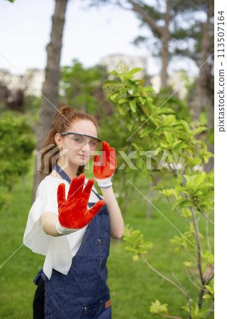 Front view of female florist standing in garden, showing hand, looking at camera. Concept of taking care of nature. Front view of female florist standing in garden, showing hand, looking at camera. Concept of taking care of nature. 113507164