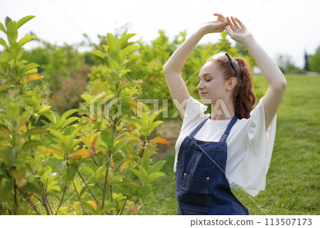 Side view of girl with red hair standing in garden, raising hands, enjoying. Concept of taking care of nature. 113507173