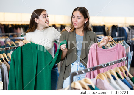 Two young women comparing green and pink cardigans while spending time together in a shopping mall 113507211