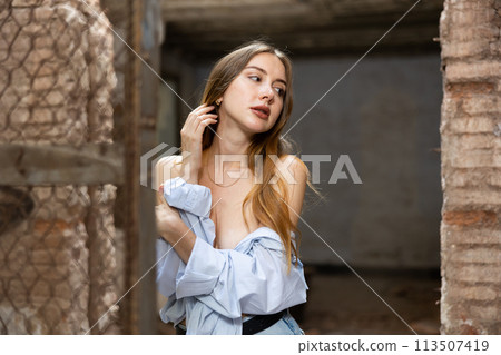 Woman in shirt pulled down from shoulders standing near wire fencing in abandoned house Woman in shirt pulled down from shoulders standing near wire fencing in abandoned house 113507419