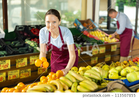 Woman selelr of vegetable department store is replenishing showcase with oranges Woman selelr of vegetable department store is replenishing showcase with oranges 113507855