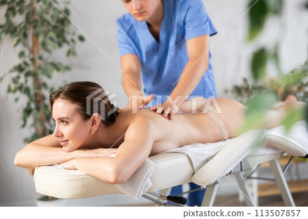 Cropped male physiotherapist in medical suit massages back of girl client Cropped male physiotherapist in medical suit massages back of girl client 113507857