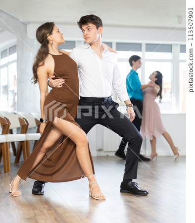 Girl and guy in festive clothes dancing slow ballroom dance during group lesson in choreography studio Girl and guy in festive clothes dancing slow ballroom dance during group lesson in choreography studio 113507901