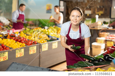 Woman seller of fruit and vegetable store works near showcase with garden-stuff, sorting cucumber Woman seller of fruit and vegetable store works near showcase with garden-stuff, sorting cucumber 113508021