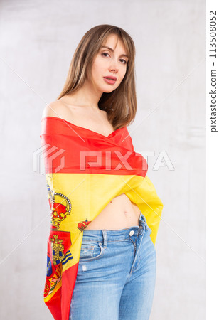 Flirting young woman posing with flag of Spain against gray wall 113508052