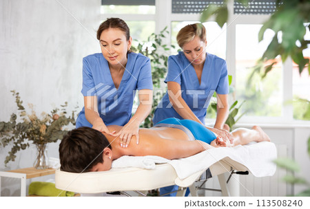 Two female masseuses perform stroking and rubbing of muscles of back and shoulders to guy client 113508240