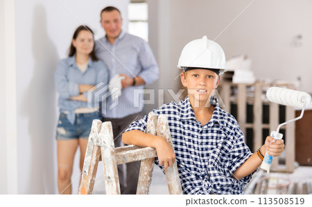 Portrait of builder boy in hard hat with a paint roller. In the background, mom and dad are renovating room Portrait of builder boy in hard hat with a paint roller. In the background, mom and dad are renovating room 113508519