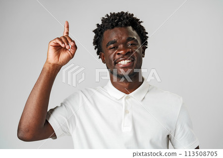 Young African Man in Casual Attire Pointing Upwards With a Smile Against a Grey Background 113508705