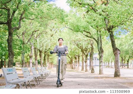 一名年輕女子拿著電動滑板在綠樹成蔭的街道上奔跑 一名年輕女子拿著電動滑板在綠樹成蔭的街道上奔跑 113508969