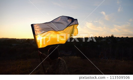 Young man in military uniform waving flag of Ukraine against beautiful sunset at background. Male ukrainian army soldier lifted national banner at countryside. Victory against russian aggression 113509506