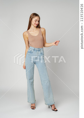 Young Woman Wearing High Rise Jeans and Tank Top posing on gray background in studio 113510416