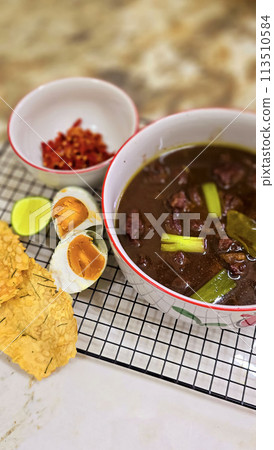 Rawon or Indonesian black beef soup, black color are from indonesian nut called kluwek. Served with lime, chili paste, salted egg, and tempe cracker. Perfect for recipe, article, or any cooking 113510584