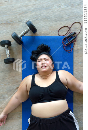 A plus-size woman lies exhausted on a blue mat, taking a breather after a vigorous workout session with dumbbell and jump rope. Top view 113511884