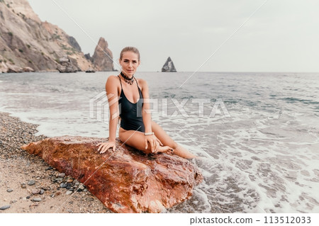 Woman travel portrait. Happy woman with long hair looking at camera and smiling. Close up portrait cute woman in black bikini posing on a red volcanic rock on the sea beach Woman travel portrait. Happy woman with long hair looking at camera and smiling. Close up portrait cute woman in black bikini posing on a red volcanic rock on the sea beach 113512033