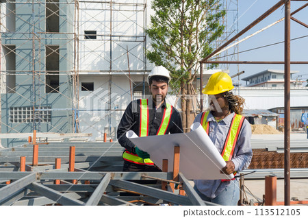 Two construction worker looking at a floor plan, standing in front of a large building site with helmet and safety gear. Day time work safety check. Work environment at the site of housing projects. Two construction worker looking at a floor plan, standing in front of a large building site with helmet and safety gear. Day time work safety check. Work environment at the site of housing projects. 113512105