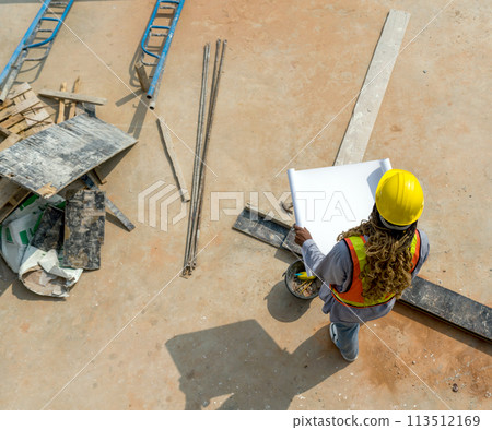 Worker in hardhat and reflective vest holding blueprint at a construction site on a sunny day. Top View. 113512169