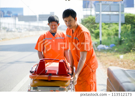 Two rescue staff in orange uniform putting a stretcher into an ambulance. Two rescue staff in orange uniform putting a stretcher into an ambulance. 113512173