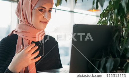 Video chat. Online work. Pretty smiling woman in hijab talking on laptop web conference digital meeting remote freelance job at cozy cafe. 113513353