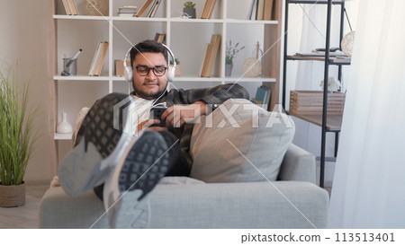 Mobile music. Enjoying song. Playlist leisure. Happy man listening to favorite tune on phone in headphones relaxing on couch in home living room. 113513401
