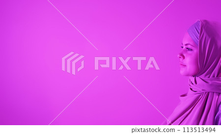 Serious profile. Islamic fashion. Confident attractive woman in elegant hijab outfit on pink neon light copy space background. Serious profile. Islamic fashion. Confident attractive woman in elegant hijab outfit on pink neon light copy space background. 113513494