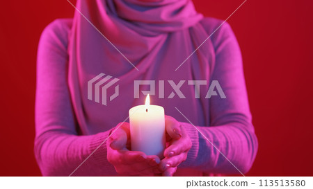 Candle pray. Ramadan night. Closeup of unrecognizable woman hands holding burning wax flame light in colorful pink neon glow on red background. 113513580