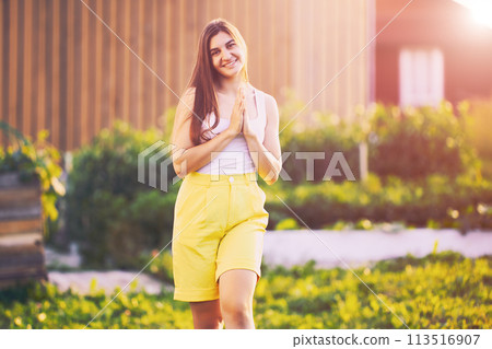 Knee length woman portrait wearing yellow bermuda shorts, young caucasian female stands against blurred rural background on summer evening before sunset. Knee length woman portrait wearing yellow bermuda shorts, young caucasian female stands against blurred rural background on summer evening before sunset. 113516907