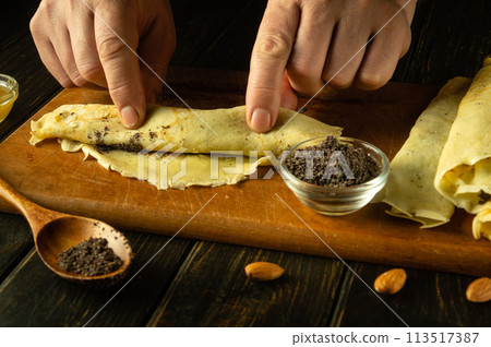 Close-up of a man hands rolling a pancake with poppy seeds on a cutting board in a home kitchen. The concept of preparing dessert for the holiday Close-up of a man hands rolling a pancake with poppy seeds on a cutting board in a home kitchen. The concept of preparing dessert for the holiday 113517387