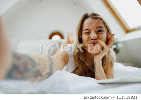 A woman is sitting comfortably on the bed, smiling and taking a selfie with her phone 113519821
