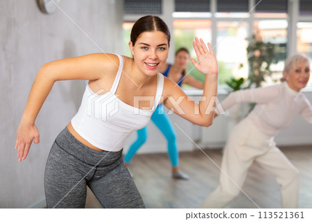 Sporty motivated young elderly women warming up with energetic dances before strength training in gym studio Sporty motivated young elderly women warming up with energetic dances before strength training in gym studio 113521361