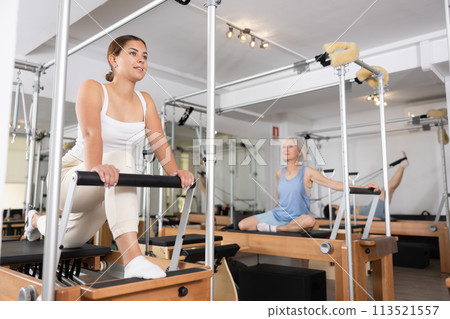 Positive young female in sportswear training twist rotation while using pilates performer bed in gym 113521557
