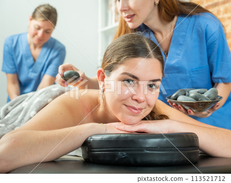 Smiling girl getting professional stone massage from two masseuse Smiling girl getting professional stone massage from two masseuse 113521712