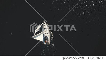 Top down of lone yacht sailing in open sea aerial. Black ocean and white sail boat at sun reflection Top down of lone yacht sailing in open sea aerial. Black ocean and white sail boat at sun reflection 113523611
