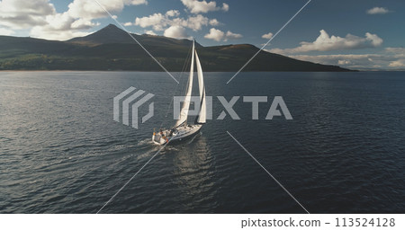 Slow motion yacht sails at mountain island shore aerial. White sailboat at Arran, Scotland, Europe Slow motion yacht sails at mountain island shore aerial. White sailboat at Arran, Scotland, Europe 113524128