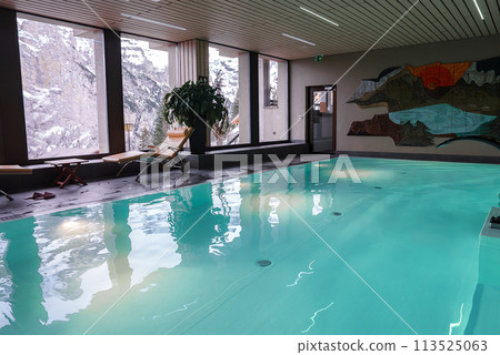 Indoor swimming pool with turquoise waters under ceiling lights, panoramic snowy mountain view, wooden lounge chairs, modern design, and colorful mural. 113525063