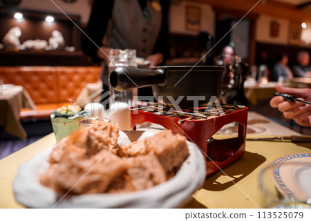 Dining scene with traditional fondue setup bread cubes on plate, black fondue pot on red stand with burner, interactive dining. Cozy restaurant ambiance, no identified location. Dining scene with traditional fondue setup bread cubes on plate, black fondue pot on red stand with burner, interactive dining. Cozy restaurant ambiance, no identified location. 113525079