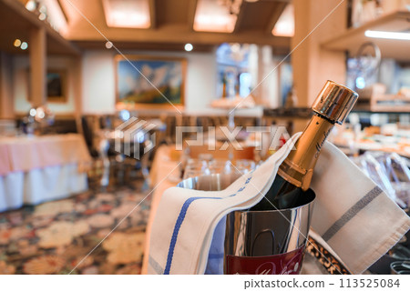 Champagne bottle in ice bucket on table with blue striped towel and patterned tablecloth in elegant restaurant dining area with warm lighting and floral carpet. 113525084