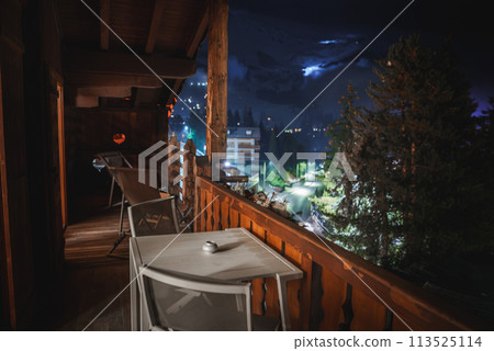 Nighttime balcony scene from mountain lodge with rustic wooden railing, white table, ashtray, and chairs. Illuminated buildings and misty mountains in the background. Perfect for a peaceful retreat. Nighttime balcony scene from mountain lodge with rustic wooden railing, white table, ashtray, and chairs. Illuminated buildings and misty mountains in the background. Perfect for a peaceful retreat. 113525114