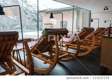 Indoor relaxation area with wooden rocking chairs lined up facing snowy mountain landscape through large glass windows. Modern, minimalistic design with residen eig sign visible. Serene atmosphere. Indoor relaxation area with wooden rocking chairs lined up facing snowy mountain landscape through large glass windows. Modern, minimalistic design with residen eig sign visible. Serene atmosphere. 113525159