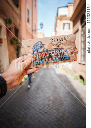 Person holds a Colosseum postcard in Rome, Italy. The background features a typical cobblestone street with buildings and a person walking. Sunny day ambiance. 113525184