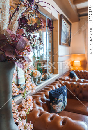 Elegant living room with large vase of artificial flowers on windowsill, leather Chesterfield sofa, framed artwork, and cozy lighting. Comfort and sophistication. Elegant living room with large vase of artificial flowers on windowsill, leather Chesterfield sofa, framed artwork, and cozy lighting. Comfort and sophistication. 113525224