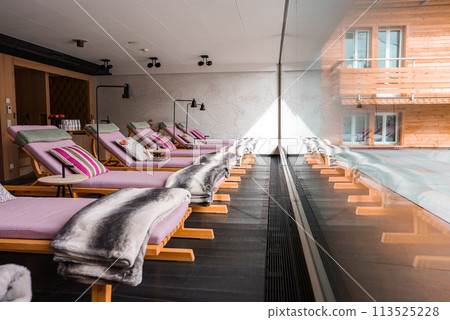 Indoor poolside relaxation area with wooden lounge chairs, purple cushions, tranquil swimming pool, large windows, minimalist design, and artistic mural. Indoor poolside relaxation area with wooden lounge chairs, purple cushions, tranquil swimming pool, large windows, minimalist design, and artistic mural. 113525228