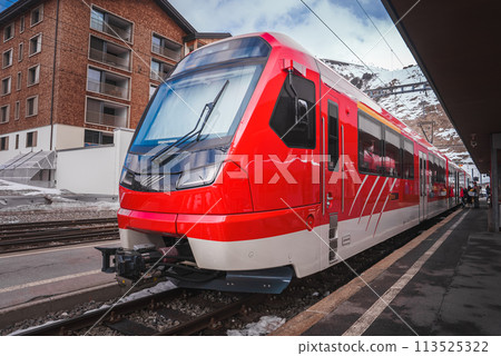 Modern red train at platform, likely part of Glacier Express in Switzerland. Sleek design with large front window for wide view. Snowy mountainous backdrop near Zermatt. 113525322
