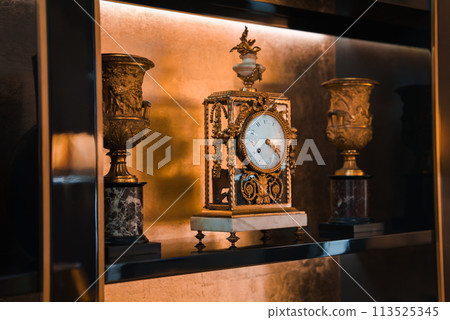 Opulent antique clock, urns with classical design, luxurious decor in Rome hotel, intricate metalwork, white clock face, elegant details, warm lighting. Opulent antique clock, urns with classical design, luxurious decor in Rome hotel, intricate metalwork, white clock face, elegant details, warm lighting. 113525345