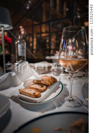 Elegant dining setting with white wine, bread, and water on a beautifully set table. Cozy, luxurious ambiance in an upscale restaurant with warm lighting. Elegant dining setting with white wine, bread, and water on a beautifully set table. Cozy, luxurious ambiance in an upscale restaurant with warm lighting. 113525443
