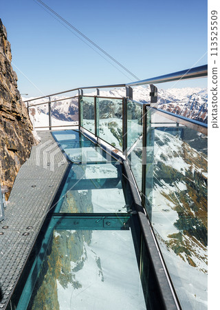 Glass floored walkway with transparent panels at Murren ski resort in Switzerland. Offers clear views of snowy landscape and dramatic cliffs. Sky is clear blue. 113525509