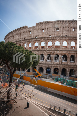Iconic Rome landmark with clear blue skies. Partial view of Colosseum shows massive stone walls towering over street and construction site with modern crane. Iconic Rome landmark with clear blue skies. Partial view of Colosseum shows massive stone walls towering over street and construction site with modern crane. 113525516