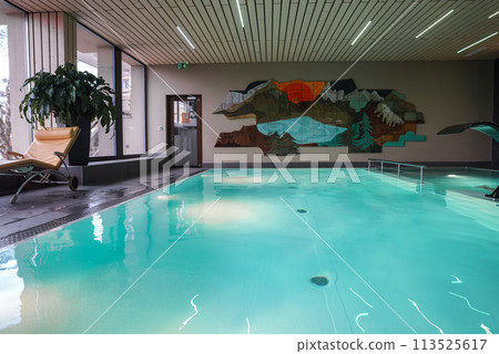 Indoor swimming pool area featuring calm turquoise waters, deck chair, potted plant, large windows, abstract wall art, wooden ceiling slats, tranquil ambiance. 113525617